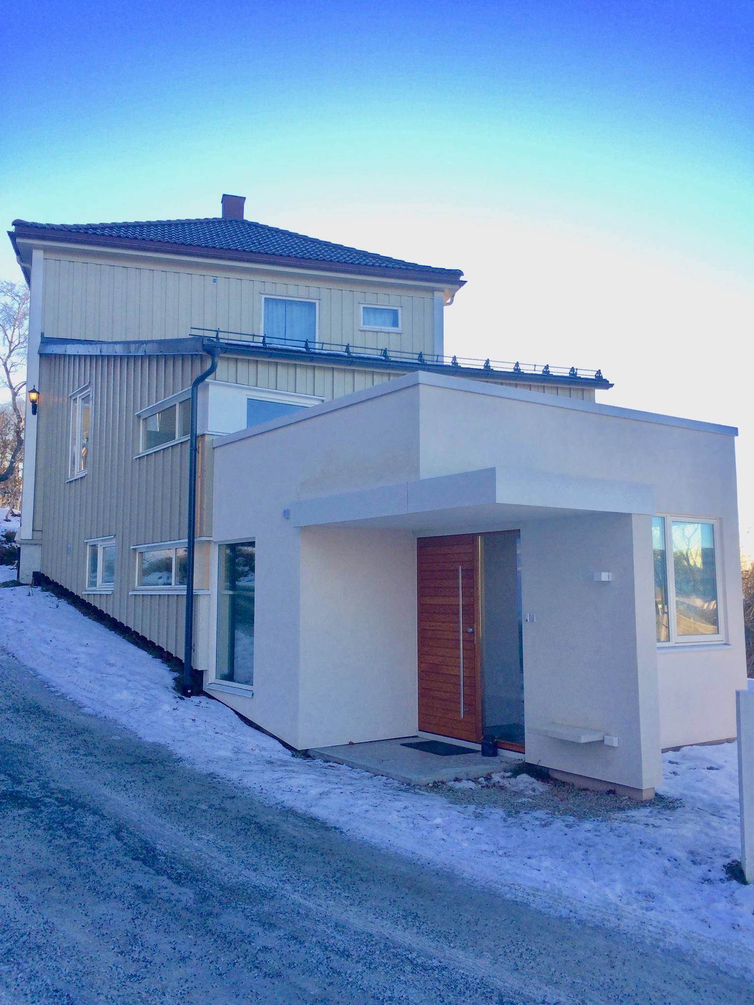 Hvitt og moderne tilbygg med inngangsparti, plassert i skrånende terreng vinterstid.