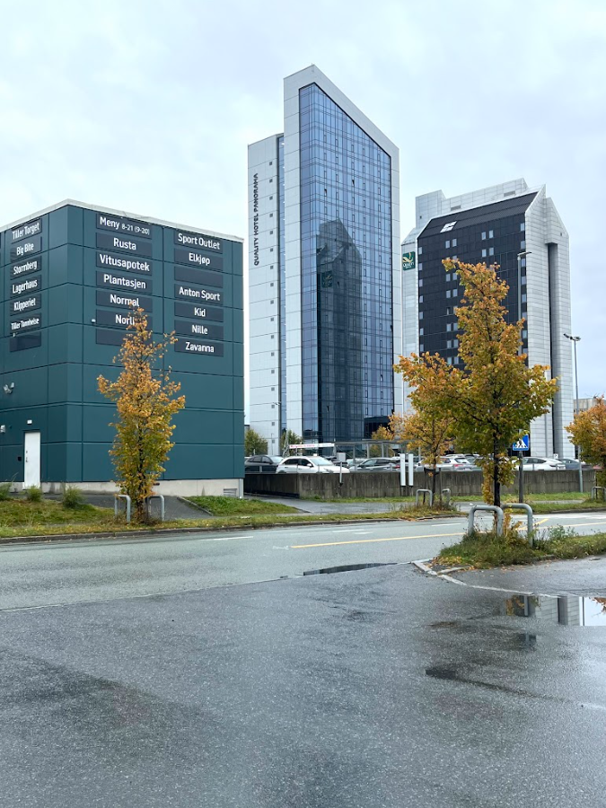 Moderne næringsbygg med glassfasader og høsttrær langs våt asfaltvei.