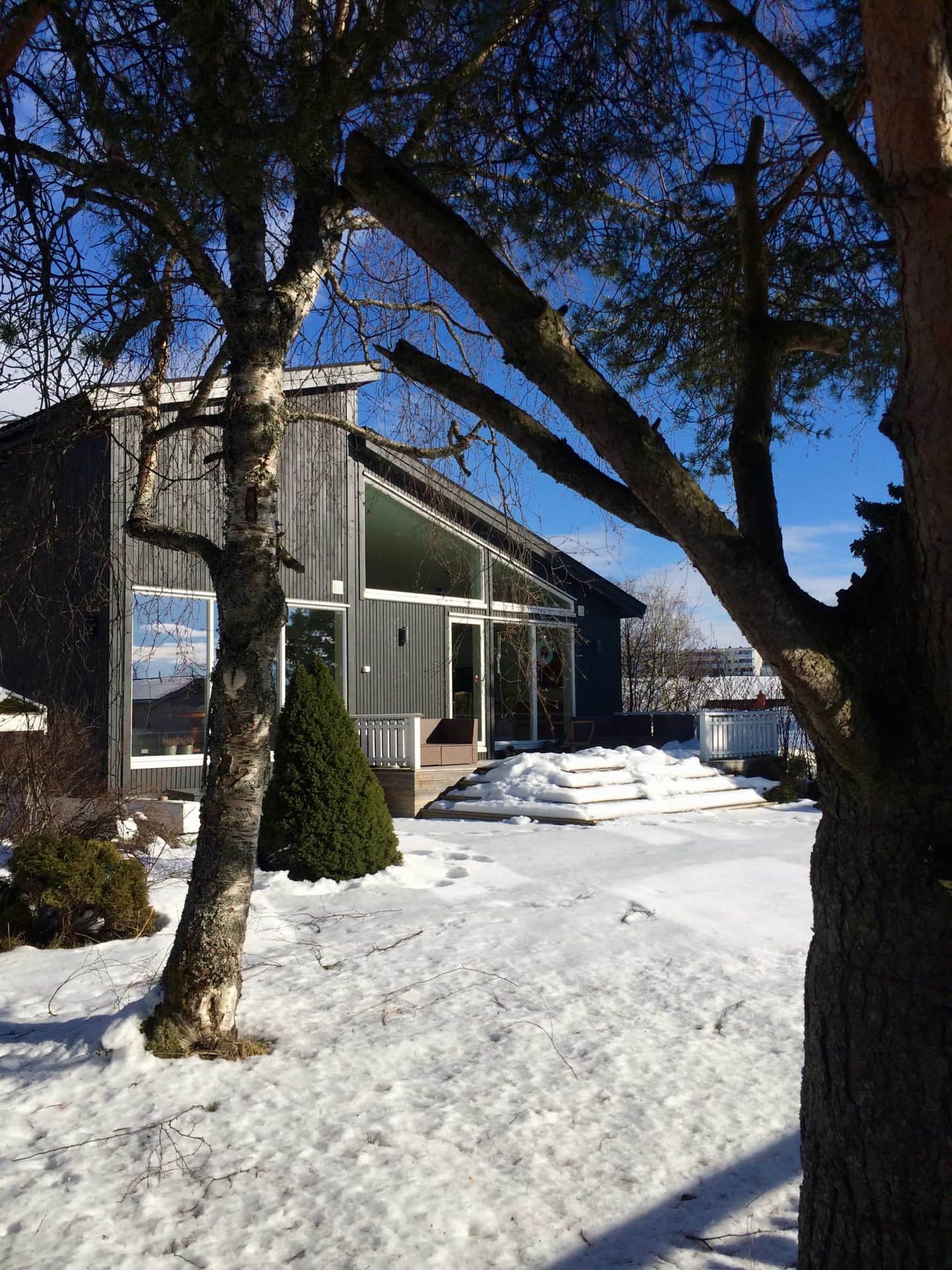 Moderne enebolig med mørk trekledning og store vinduer, omgitt av snødekt hage og trær en solrik vinterdag.
