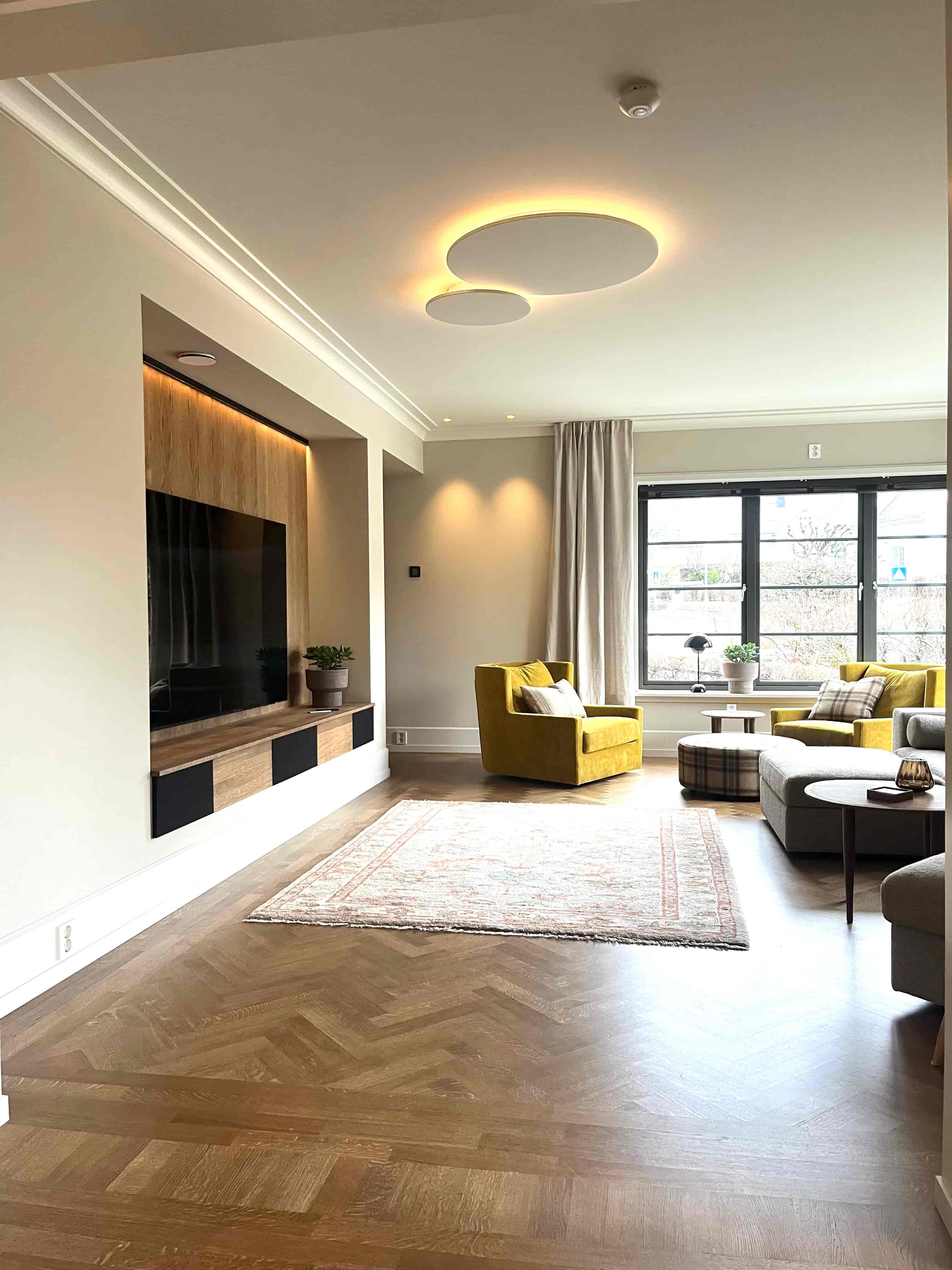 Moderne stue med fiskebeinsparkett, innebygd TV-vegg i tre, gule lenestoler og store vinduer med gardiner.